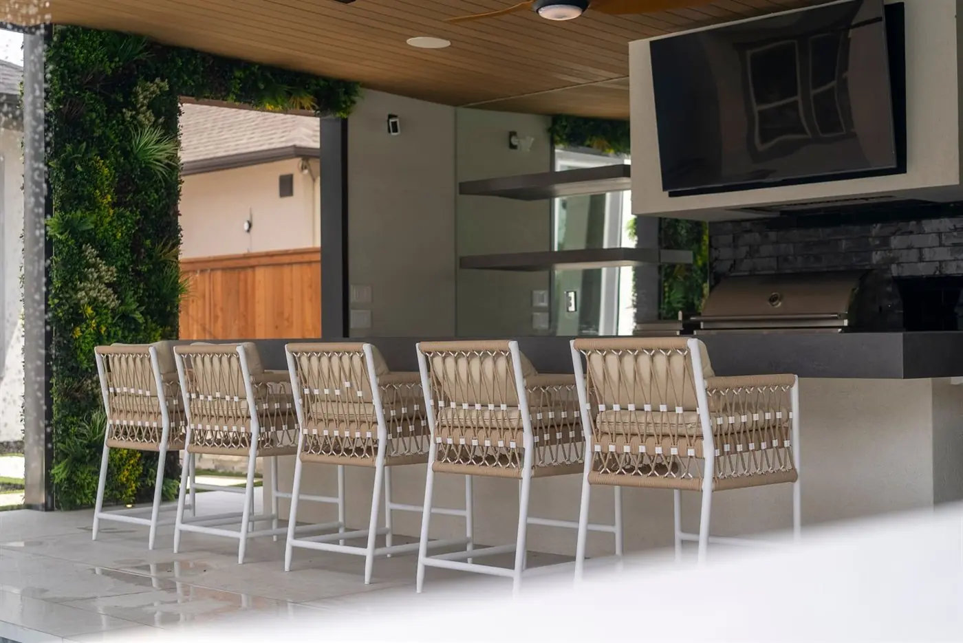 Outdoor bar counter with woven stools beside a grill and wall-mounted TV