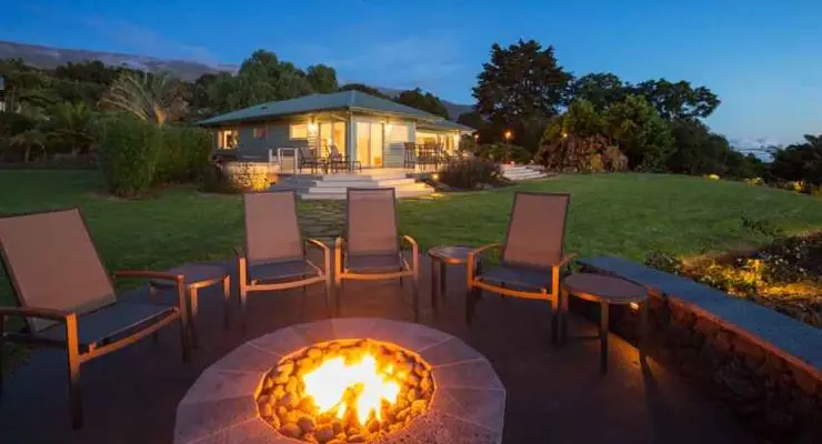 Stone fire bowl with chairs around it in front of a lit house at dusk